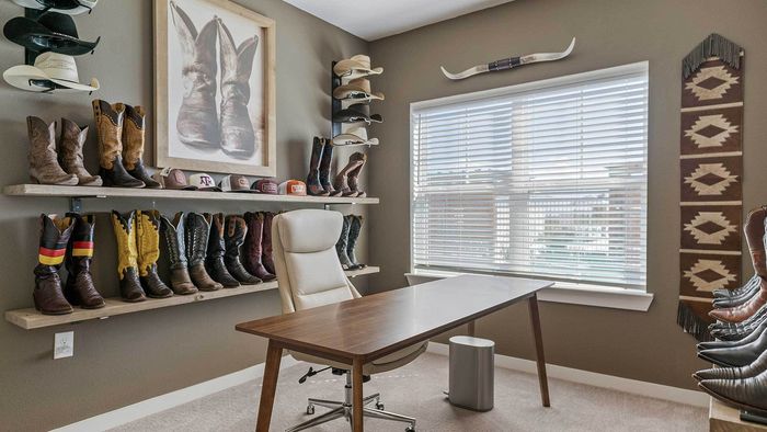 Apartment home office setup featuring a wooden desk, office chair, a large window with blinds, and custom shelving displaying boots and hats against a grey wall.