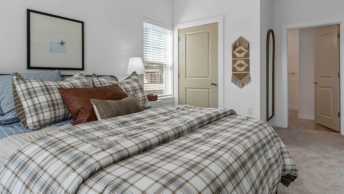 Cozy apartment bedroom featuring a large bed with plaid bedding, a window with blinds, and an en-suite doorway leading to a walk-in closet and bathroom.