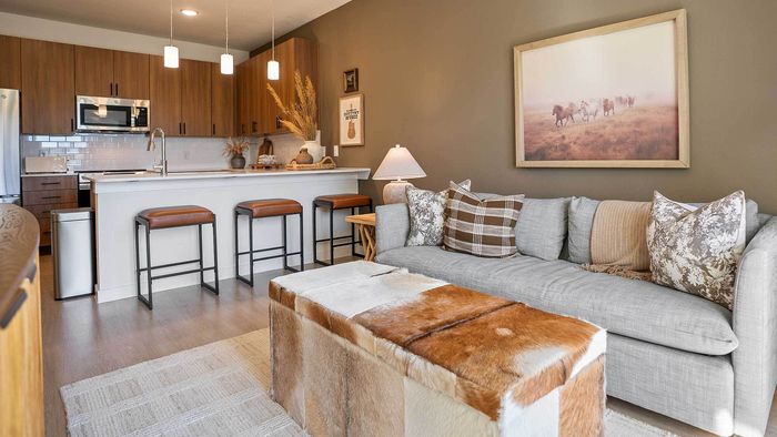 Open-concept apartment interior showing a modern kitchen with a large island and barstools flowing seamlessly into a stylish living room with a grey sofa.