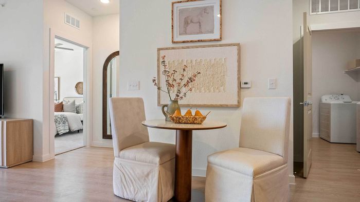 Cozy apartment dining nook featuring a small round wooden table and two upholstered chairs, with doorways leading to a bedroom and a dedicated laundry room.