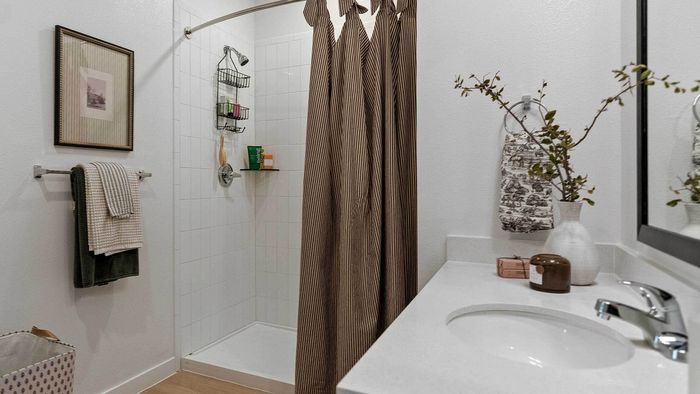Close-up of a modern apartment bathroom featuring a white quartz vanity with a chrome faucet and undermount sink, alongside a walk-in shower with white tile.