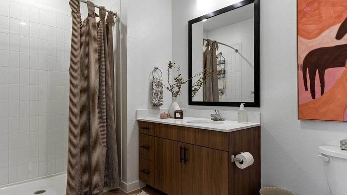 Interior view of a contemporary apartment bathroom showcasing a wood-grain vanity cabinet with dark hardware, white quartz countertops, a framed mirror, and vibrant wall art.