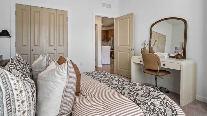 Cozy apartment bedroom featuring a bed with patterned linens, a dedicated modern vanity desk space with a mirror, closet space, and an open doorway leading to the kitchen.