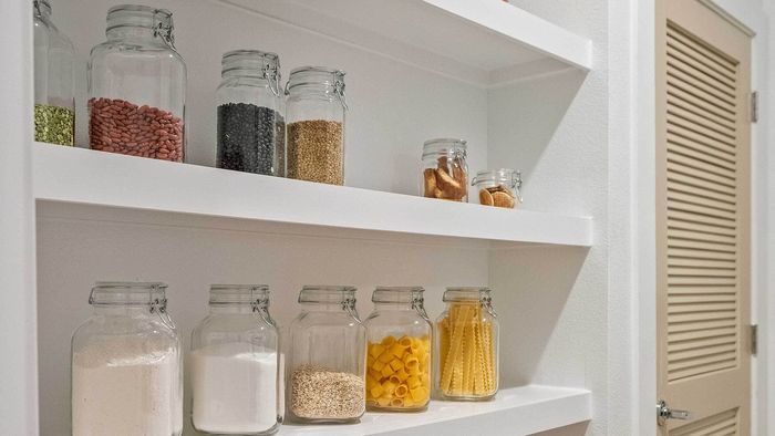 Built-in white pantry shelving in an apartment, beautifully organized with clear glass jars filled with dry goods, located next to a closed louvered door.