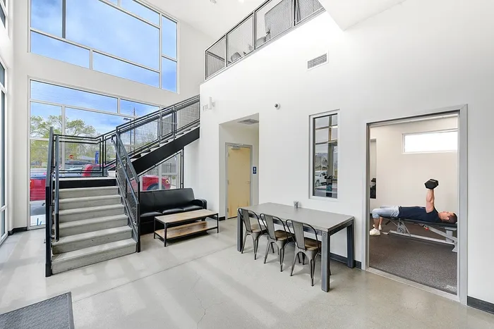 Modern apartment interior showcasing a spacious open-plan living area with high ceilings, large windows, a sleek staircase, and a small fitness room.