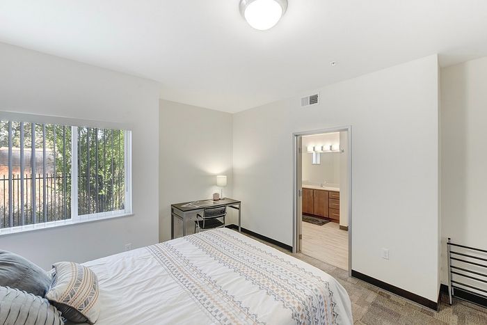 Bright and spacious bedroom featuring a cozy bed, modern desk, and sunny window view, leading to an ensuite bathroom with elegant fixtures.