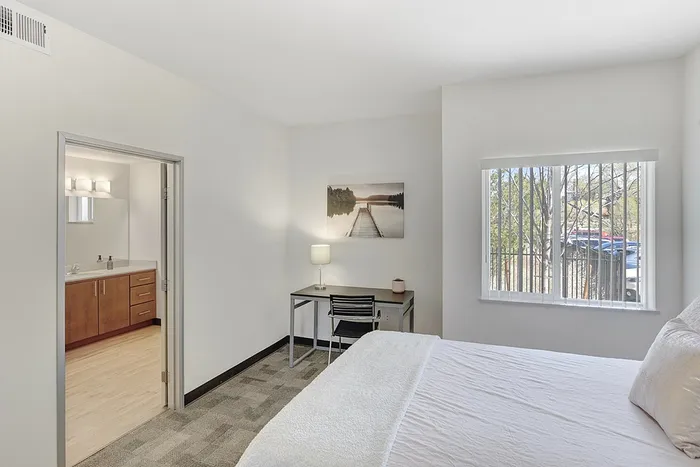 Bright, inviting bedroom with a cozy bed, modern desk, and a scenic view through large windows, featuring an ensuite bathroom with a stylish vanity.