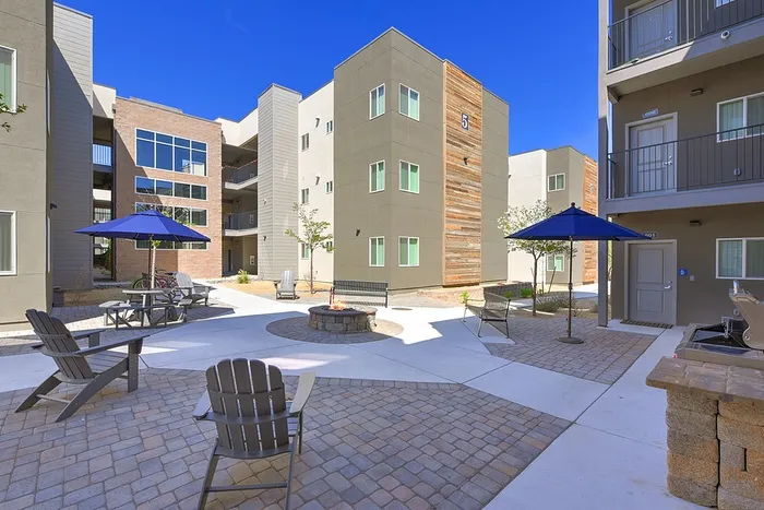 Modern apartment complex courtyard with stylish seating, vibrant blue umbrellas, and a cozy fire pit under a clear sky.