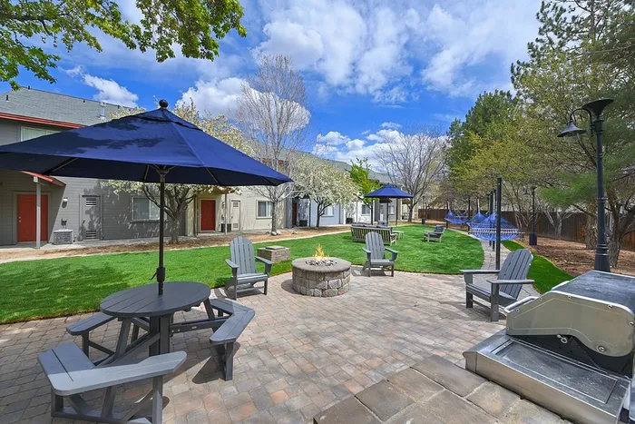 Charming outdoor patio featuring cozy fire pit, elegant seating under blue umbrellas, and lush greenery, ideal for relaxation and gatherings.