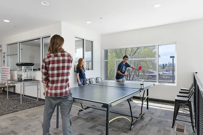 Bright and inviting recreational room with people enjoying a game of ping pong, featuring large windows and ample seating.