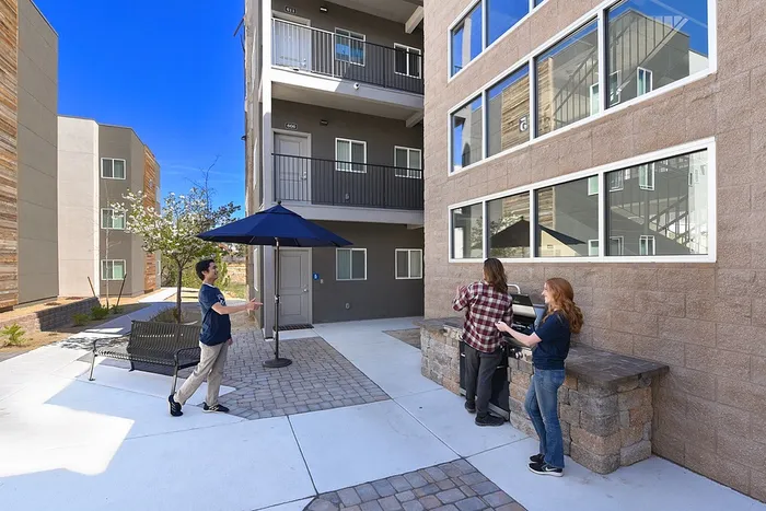 Vibrant apartment courtyard with people enjoying a barbecue under a blue umbrella, surrounded by modern architecture.