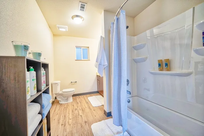 Bright and modern bathroom with elegant fixtures, featuring wood flooring, a spacious bathtub with shower, and stylish shelving for a touch of luxury.