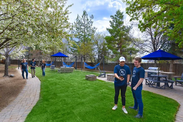 Vibrant community garden featuring lush green grass, blooming trees, and comfortable seating. Residents enjoy a sunny day with hammocks and picnic tables under blue umbrellas, perfect for relaxation and socializing.