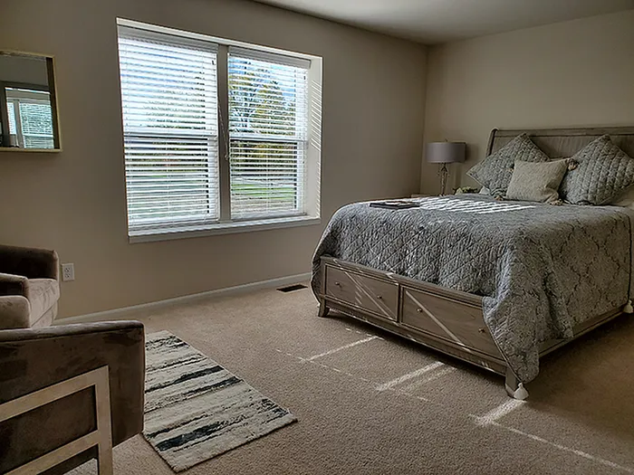 Inviting bedroom with a cozy quilted bed, abundant natural light from large windows, and a plush armchair in a serene setting.