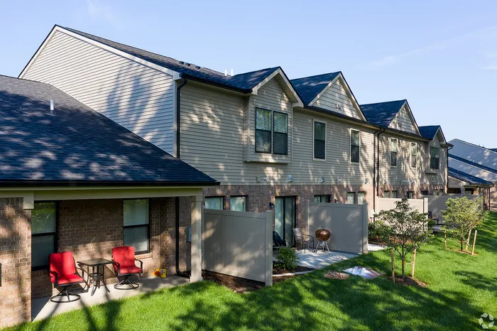 Charming townhouse exterior with a modern brick finish, featuring private patios with red chairs, perfect for relaxing amid lush green landscaping.