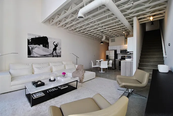 Modern open-concept living space featuring a sleek white sofa, stylish armchairs, and contemporary art. Spacious kitchen area with stainless steel appliances and elegant dining setup, enhanced by industrial-style ceiling beams.