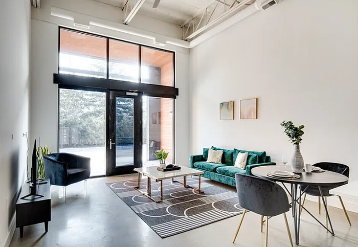 Bright and modern living space with high ceilings, large windows, and chic turquoise sofa. Contemporary dining area and artistic decor enhance the inviting ambiance.