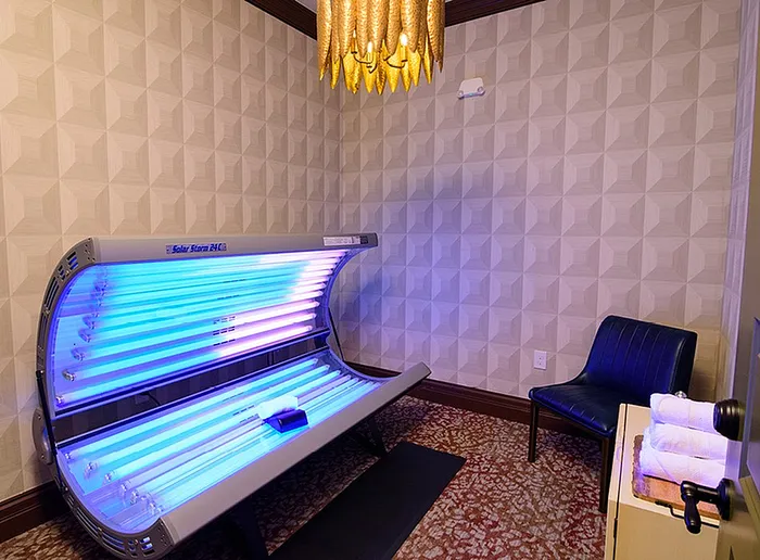 Luxurious room featuring a modern tanning bed with glowing lights, plush seating, elegant patterned walls, and a stylish chandelier.