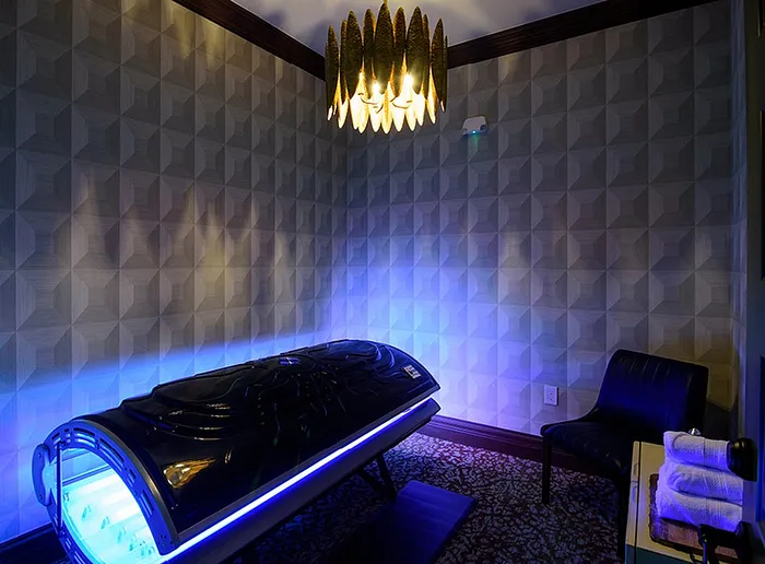 Chic wellness room featuring a sleek tanning bed under a stylish chandelier, with a cozy seating area and luxurious decor.