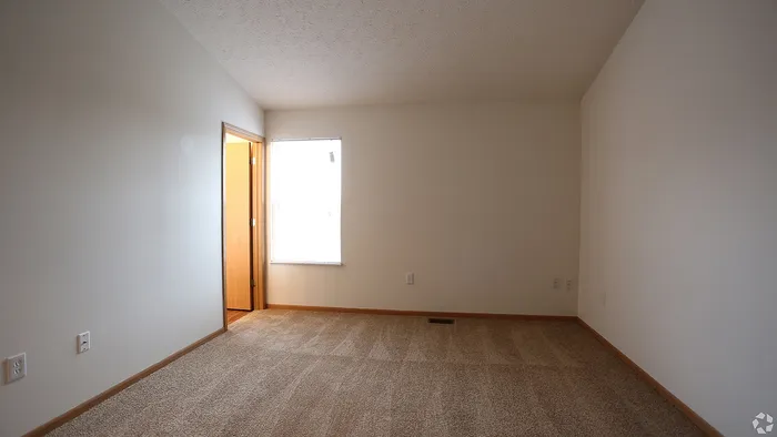 Bright and inviting bedroom with plush carpeting, large window for natural light, and neutral walls for a modern touch.