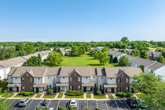 Aerial view of a charming residential neighborhood featuring picturesque townhouses, lush greenery, and ample parking space under a clear blue sky.