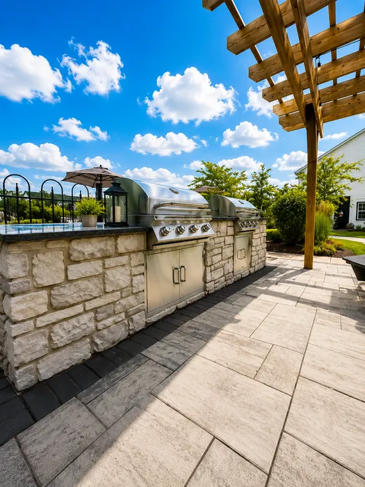Chic outdoor kitchen with sleek stainless steel grills, rustic stone countertop, and modern pergola under a vibrant blue sky.