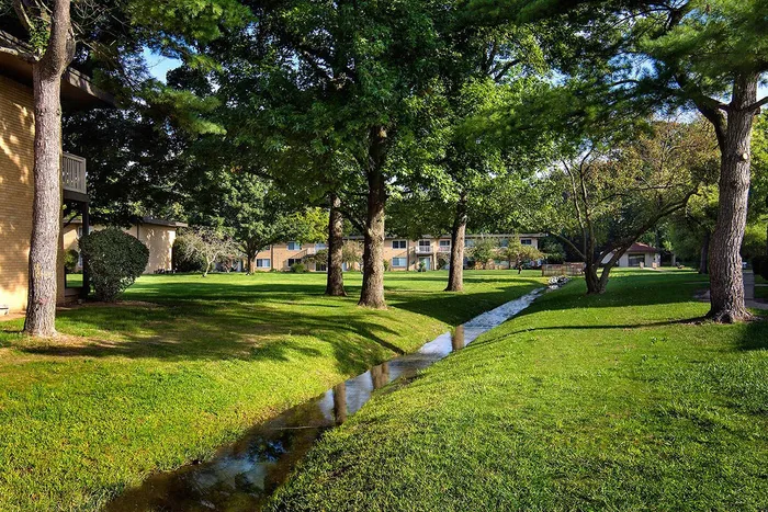 Serene apartment complex surrounded by lush green lawns and tall trees, featuring a charming stream and inviting outdoor spaces.