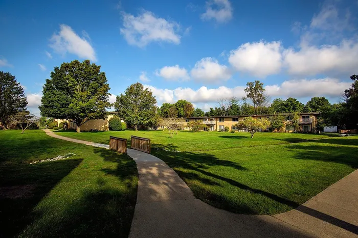 Scenic apartment complex with lush green lawns, mature trees, and a charming walkway under a clear blue sky.