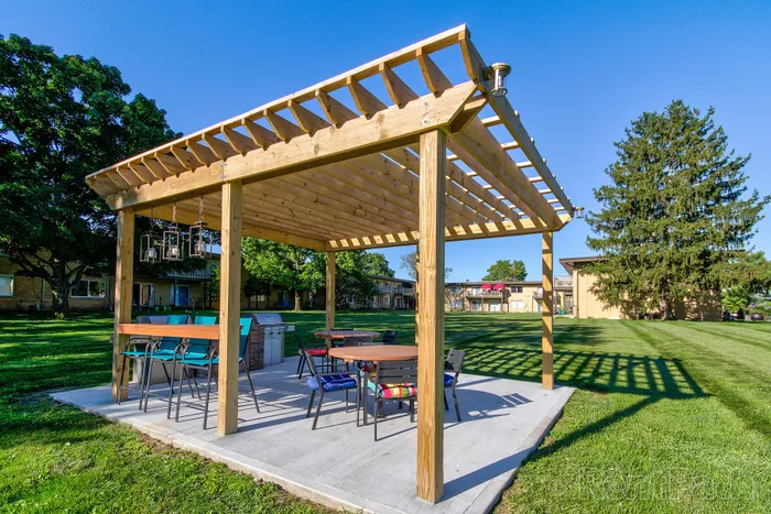 Charming outdoor pavilion with modern seating and a barbecue grill, set in a lush green community area under a clear blue sky.
