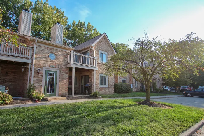 Charming brick apartment featuring a welcoming front porch, lush greenery, and a serene suburban setting.