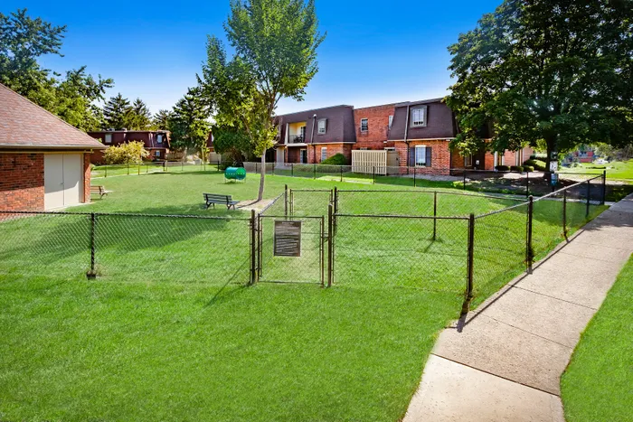 Charming apartment complex with lush green lawn, fenced area, and inviting benches under the shade of mature trees.