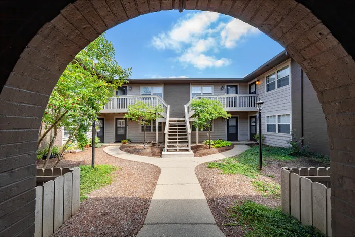 Charming apartment building with a landscaped courtyard, featuring a central staircase and elegant archway entrance under a bright blue sky.