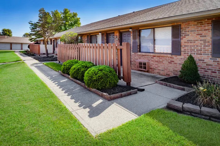 Charming brick apartment exterior with a cozy front patio, lush greenery, and vibrant landscaping.
