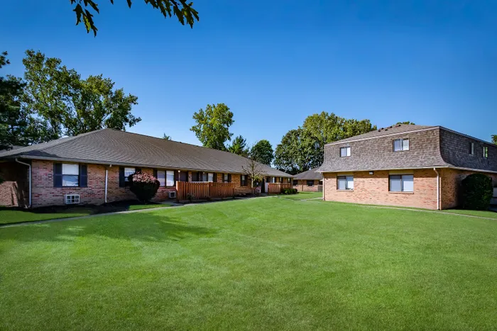 Charming brick apartment buildings surrounded by lush green lawns and mature trees basking in bright sunshine.