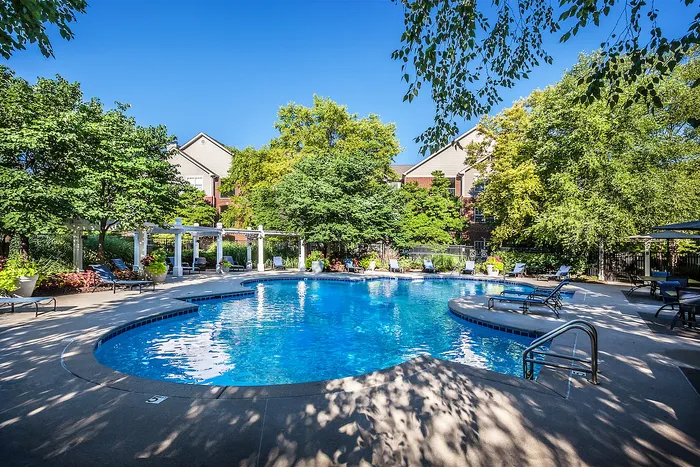 Inviting outdoor swimming pool area surrounded by lush greenery and stylish loungers, set against charming residential buildings under a clear blue sky.