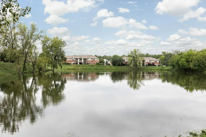 Scenic view of peaceful apartment complex nestled by a tranquil lake surrounded by lush greenery under a bright blue sky.