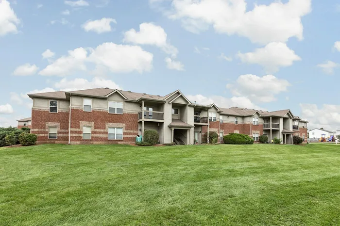Charming apartment complex with brick facade, lush green lawns, and spacious balconies under a bright blue sky.