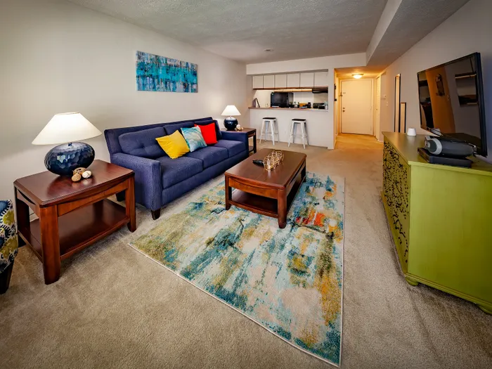 Inviting living room featuring a plush blue sofa with colorful pillows, modern decor, and a stylish open kitchen. Warm lighting and artistic rug enhance the charming ambiance.