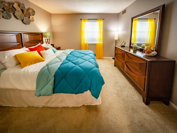 Inviting bedroom featuring a cozy bed with vibrant pillows, elegant wooden furniture, and sunlight streaming through yellow-accented windows.