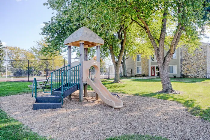 Charming playground area with a slide nestled among lush trees, adjacent to a modern apartment building, perfect for family living.