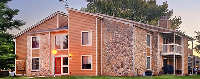 Charming two-story apartment building with a rustic stone facade and warm wood paneling, set amidst lush greenery and vibrant landscaping.