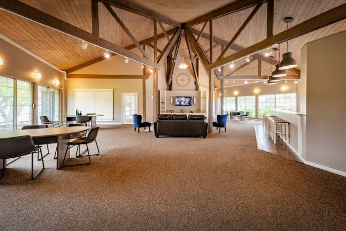 Elegant and spacious open-concept living area with high exposed beam ceilings, ambient lighting, and large windows offering lush garden views. Features cozy seating arrangements and a sleek bar area perfect for entertaining.
