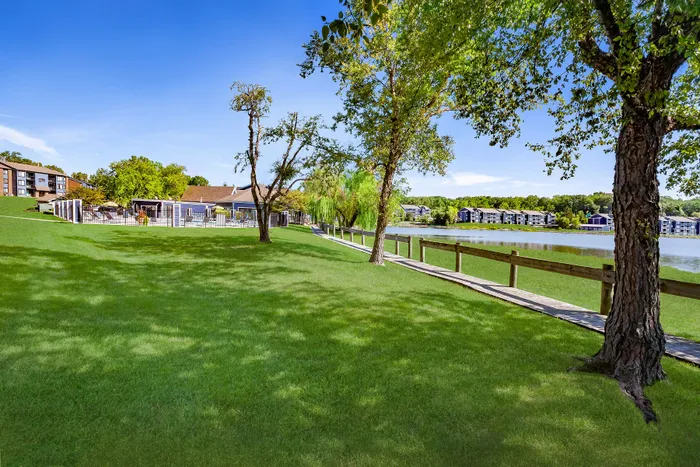 Serene lakeside apartment view featuring lush green lawns, mature trees, and a scenic walkway.