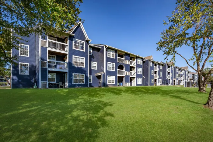 Charming three-story apartment building with stylish blue facade and inviting balconies, surrounded by lush green lawn and vibrant trees, against a clear blue sky.