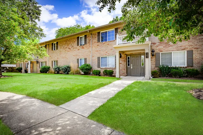 Charming brick apartment building with lush green landscaping and a welcoming front entrance, perfect for comfortable living.