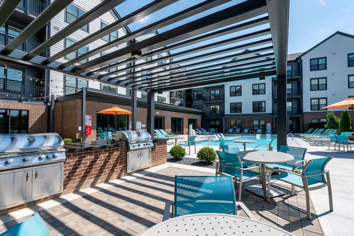 Modern apartment complex featuring a stylish outdoor patio with stainless steel grills, vibrant turquoise seating, and a sparkling swimming pool under a sunny sky.