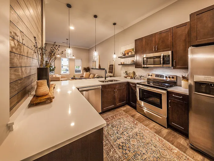 Inviting apartment kitchen with warm wooden cabinetry, modern stainless steel appliances, and elegant pendant lighting, opening to a cozy living space with ambient natural light.