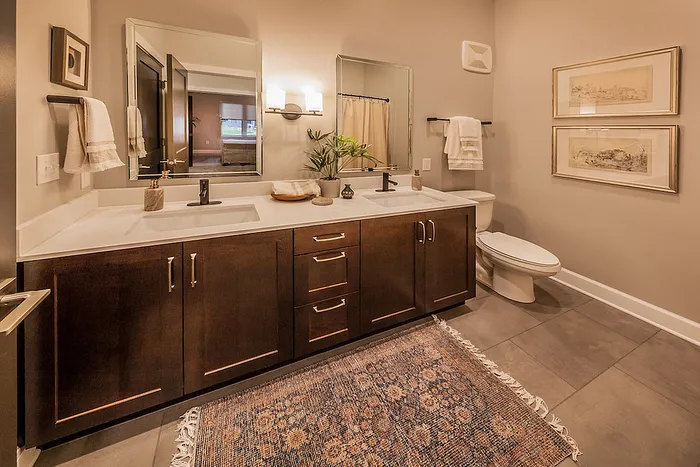 Elegant bathroom featuring a sophisticated double vanity with dark wood cabinets, warm lighting, and modern fixtures, enhanced by decorative wall art and a plush patterned rug.