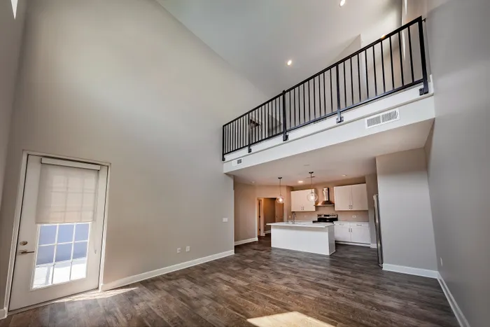 Modern loft apartment featuring high ceilings, elegant railing, stylish kitchen with pendant lighting, and sunlit door accenting the open floor plan.