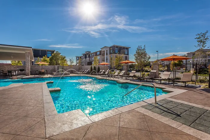 Luxurious apartment community featuring a sunlit pool with elegant loungers, water fountains, and vibrant orange umbrellas, surrounded by modern landscaping and contemporary buildings.